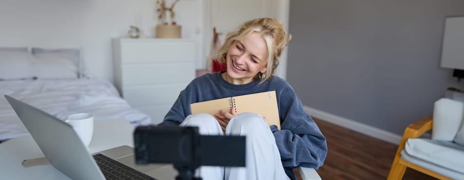 Social media and lifestyle content concept. Young happy smiling woman, sits in her room with notebook, talks at digital camera, having conversation with followers, does lifestyle vlog.