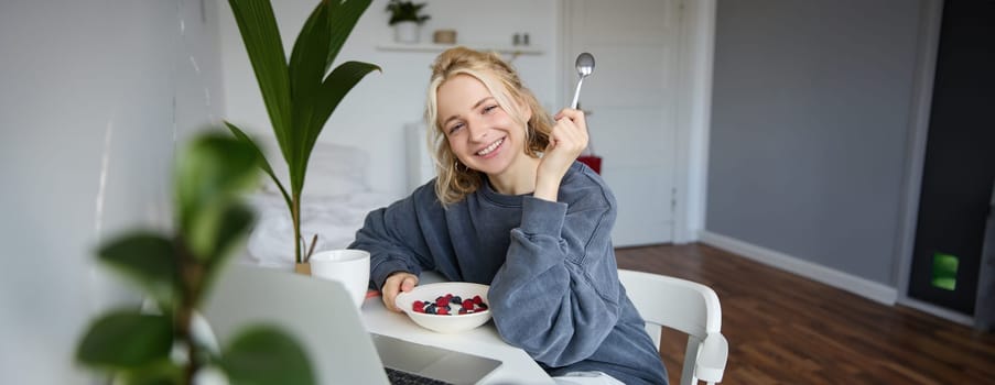 Portrait of smiling blond young woman, eating in front of laptop, watching videos online while having breakfast, enjoying dessert, sitting in bedroom.
