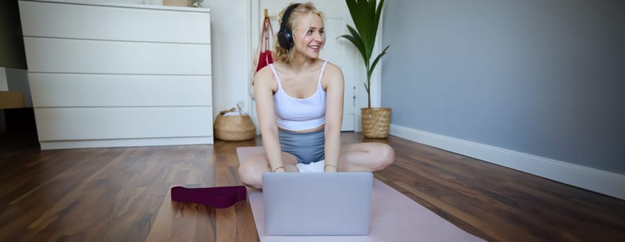 Portrait of smiling sporty woman at home, doing workout on rubber yoga mat, using laptop and wireless headphones.