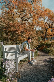 a teenager sits on a bench in the park drinks coffee from a thermo mug and looks into a phone. Portrait of handsome cheerful guy sitting on bench fresh air using device browsing media smm drinking latte urban outside outdoor.