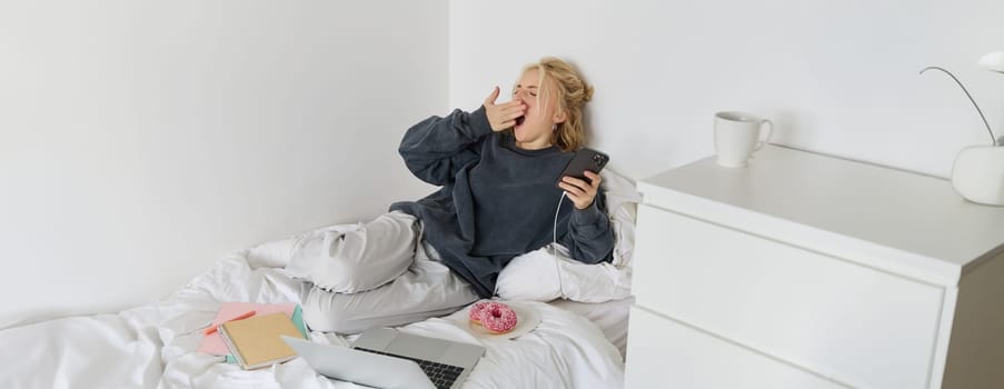 Portrait of young woman spending lazy weekend at home, lying in bed with smartphone and laptop, eats doughnut and yawns, feels sleepy or bored.