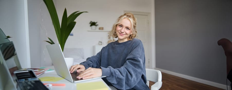 Portrait of young smiling beautiful woman, student studying remotely in her room, e-learning, using laptop, sitting on chair in bedroom, looking happy at camera.