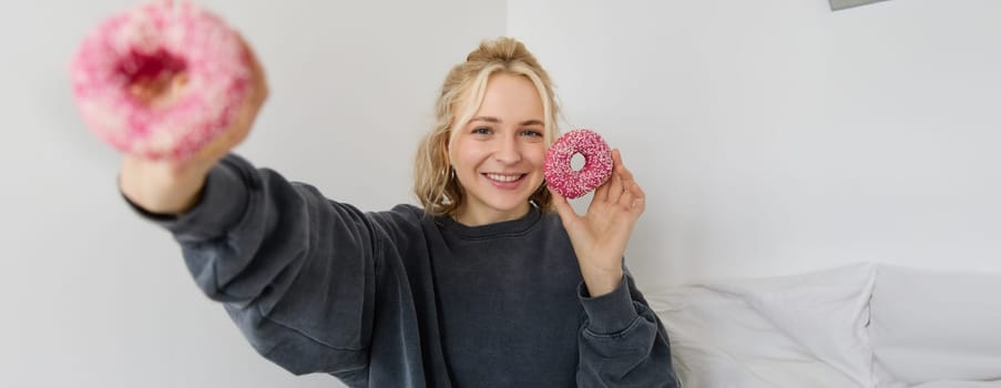 Portrait of beautiful blond woman, showing two pink doughnuts, delicious pastry, eating dessert and smiling, sharing food with you.