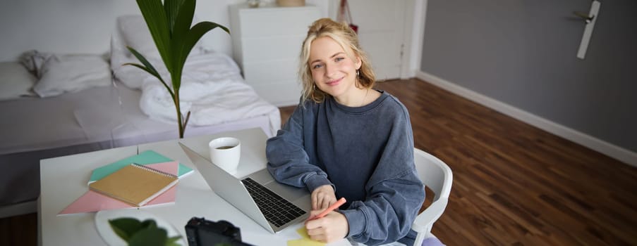 Portrait of smiling young woman, female student, doing distance learning course, using laptop, studying at home, writing down, making notes.
