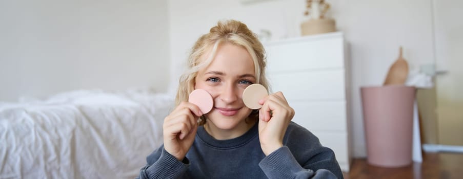 Image of young blond woman, girl records video about makeup, shows skin tone beauty products, sits in a room on floor.