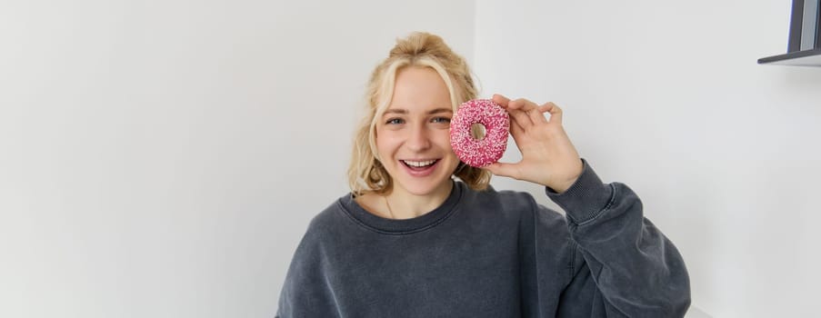 Close up portrait of cute young blond woman, showing pink doughnut near her face, smiling and looking happy. People concept