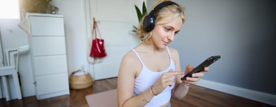 Close up portrait of blond young woman choosing fitness podcast, looking at her mobile phone, checking workout app on smartphone, wearing wireless headphones.