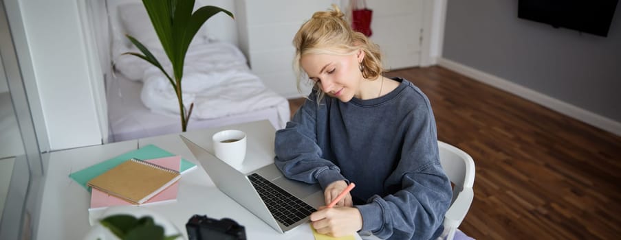 Portrait of smiling young woman, female student, doing distance learning course, using laptop, studying at home, writing down, making notes.