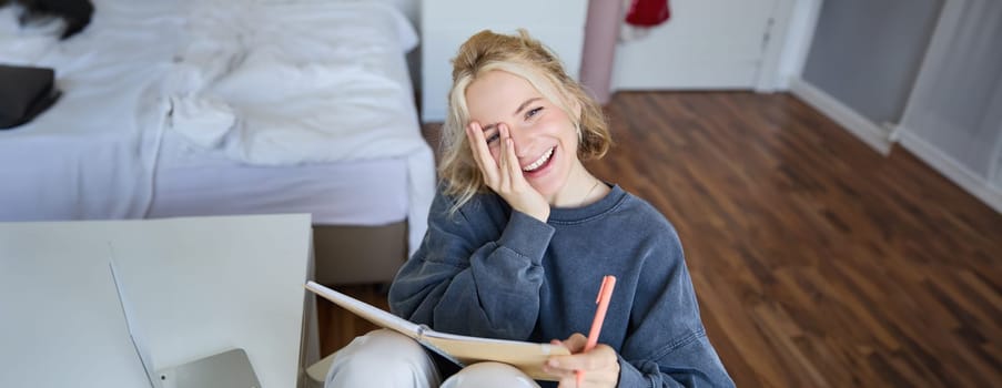 Portrait of happy, young blond woman, sitting in bedroom with notebook and pen, laughing and smiling, writing in journal, doing homework and smiling.