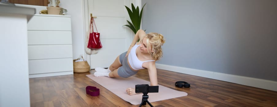 Portrait of young woman, fitness instructor recording video of her standing in a side plank, doing workout at home on camera, creating content for social media.