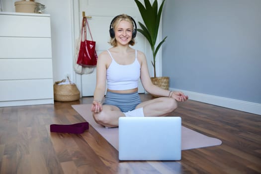 Portrait of fit and healthy woman at home, practice yoga, sitting on rubber mat, listening to instructions online, using meditation music to relax, following guidance on laptop.