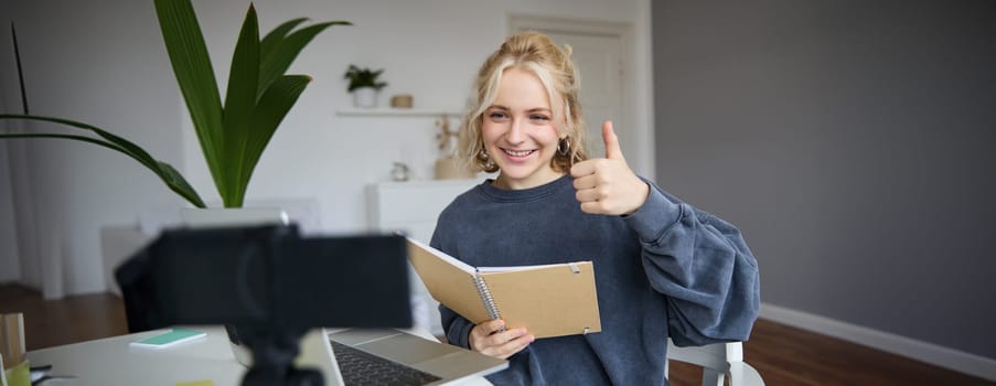 Portrait of smiling blond woman, social media vlogger, reads notes from notebook and looks at digital camera, records video in her room, shows thumbs up.