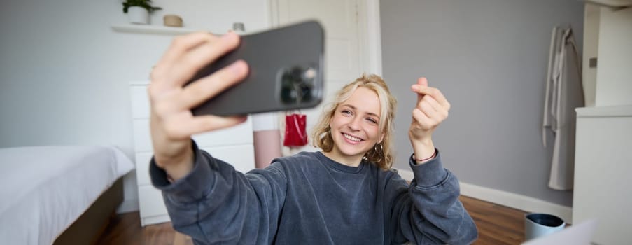 Image of young woman, vlogger taking selfie in her room, talking to her followers during online live stream, using smartphone app to chat with audience, smiling and looking happy.