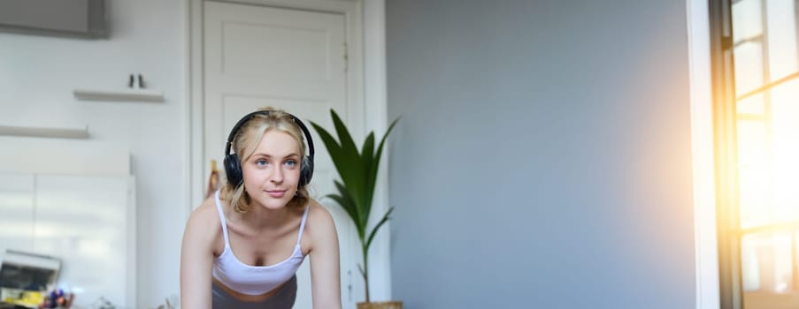 Portrait of fit and healthy woman doing workout at home, standing in plank in wireless headphones, listens to fitness instructor online, using laptop to watch sport videos.
