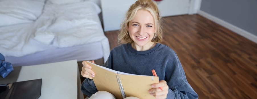 Close up portrait of beautiful smiling blond girl, holding notebook, making notes in a journal or diary, writing her thoughts, looking happy at camera.