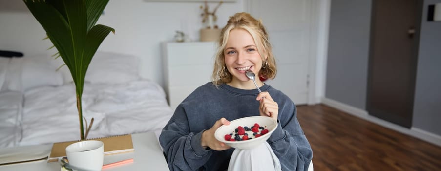 Portrait of smiling blond woman, eating breakfast, holding bowl and spoon, sitting in bedroom, looking happy at camera. Lifestyle concept