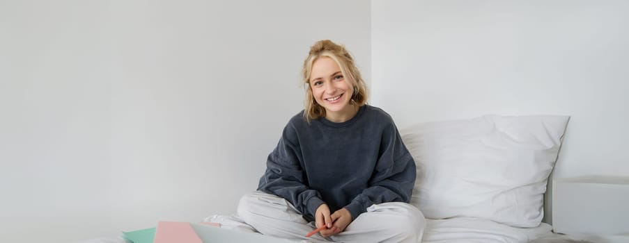 Portrait of young beautiful woman sitting on bed with laptop and notebooks, working from home, freelancing. Female student studying online, video chats, connects to lesson remotely.