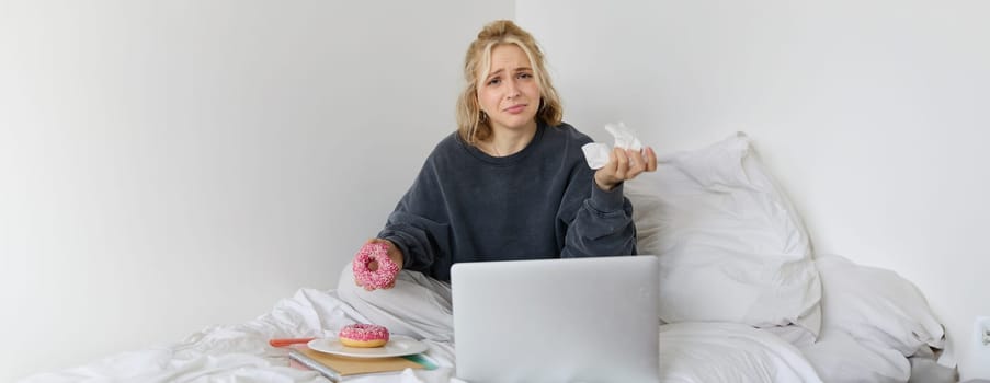 Portrait of disappointed, depressed young woman, crying, sitting on bed with laptop, eating comfort food, holding doughnut and wiping tears off.