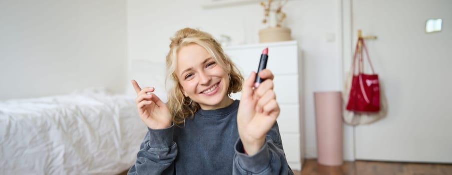 Portrait of smiling beautiful woman in her room, sitting and showing lipstick, recommending favourite beauty product, content maker recording a video of herself for social media blog.