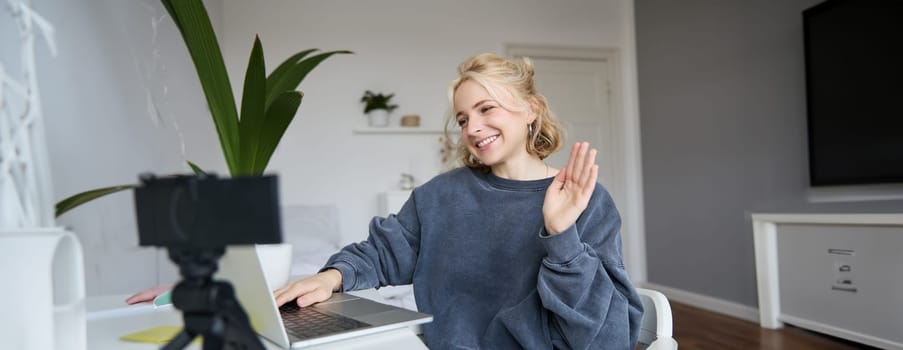 Image of smiling beautiful young woman, chats online on laptop, waves hand, says hello, records video on digital camera, vlogging, creates content for her blog.