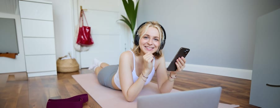 Portrait of young woman workout, watching exercise videos on laptop in headphones, lying on rubber mat with mobile phone and smiling.