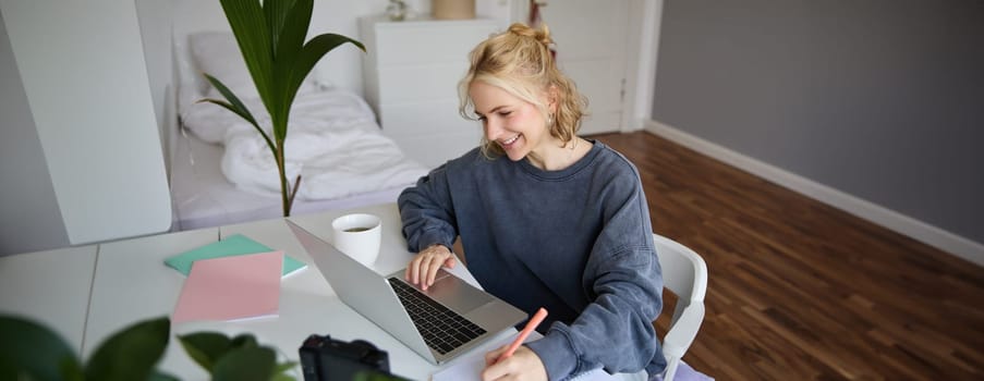 Portrait of smiling young woman, female student, doing distance learning course, using laptop, studying at home, writing down, making notes.