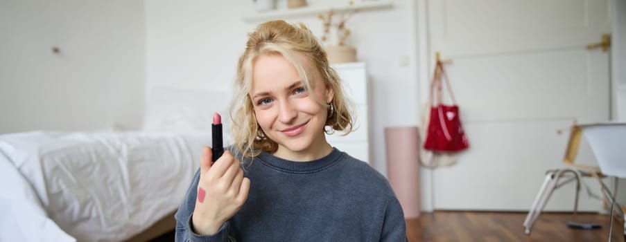 Portrait of smiling blond woman, recording video, online live stream about beauty products, content maker rates her makeup products, shows lipstick on camera.
