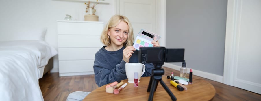 Lifestyle blogger, records video in her room, has a camera on coffee table, shows eyeshadow palette to her followers, does makeup tutorial, vlogger working indoors, creates content for social media.