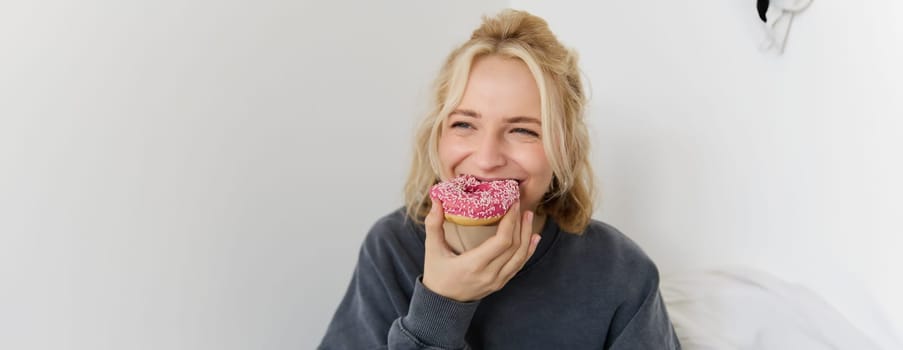 Close up portrait of happy, cute blond woman, holding doughnut, eating sweet, delicious comfort food, showing dessert at camera. Food concept