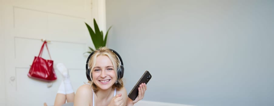 Vertical shot of young blond woman in headphones, lying on rubber mat, using laptop, holding mobile phone, listens to music or workout podcast, doing exercises at home.