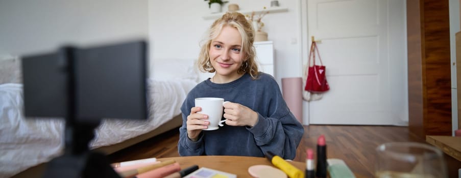 Portrait of cute blond woman, video content maker, vlogger recording on digital camera in bedroom, drinking cup of tea and talking to audience.