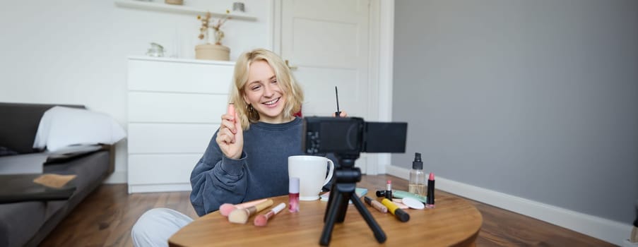 Image of cheerful, beautiful young lifestyle blogger, woman sitting on floor and recording video about makeup, holding mascara, making lifestyle content for her social media account and followers.