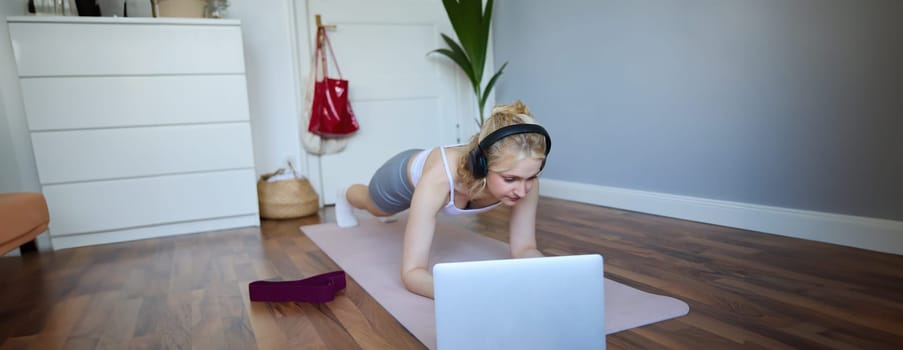 Portrait of young athletic woman in wireless headphones, standing in plank, following online workout video, using laptop to join training session on the internet.
