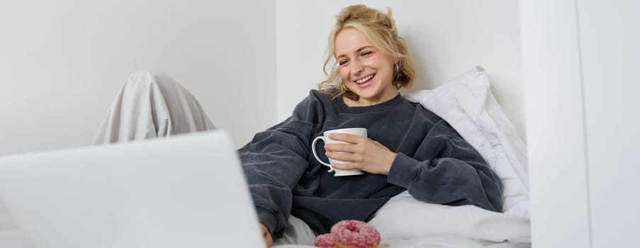 Portrait of candid, happy young woman lying in bed, looking at laptop screen, holding cup of tea and eating doughnut, staying at home on weekend, spending quite time alone.