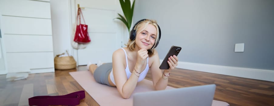 Portrait of young fitness woman, choosing workout on laptop, wearing headphones and using mobile phone exercise app. Sport and lifestyle concept