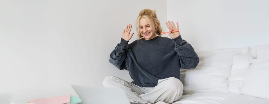 Portrait of young smiling woman working from her bed, sitting in bedroom with laptop and documents, waving hands during video chat, tutor greets her student via internet.