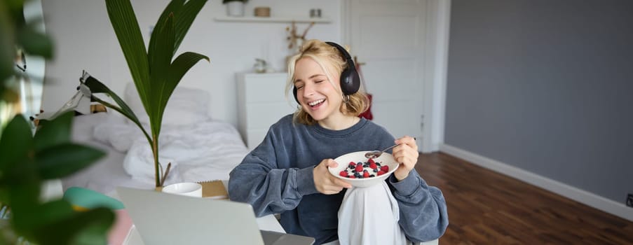 Portrait of smiling young woman, watching tv show in headphones, eating breakfast and looking at laptop screen.