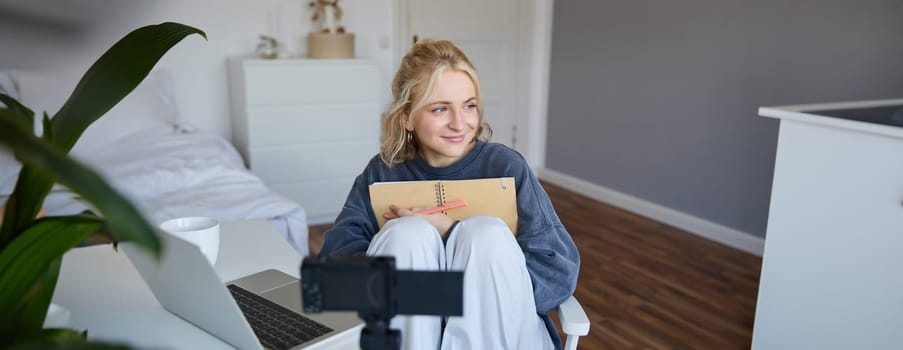 Social media and lifestyle content concept. Young happy smiling woman, sits in her room with notebook, talks at digital camera, having conversation with followers, does lifestyle vlog.
