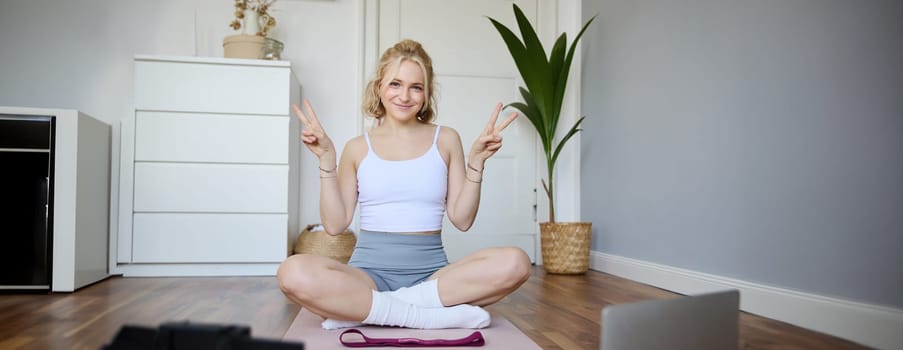 Portrait of cute blond woman shows peace sign, smiles, records yoga vlog on digital camera, workout at home on rubber mat in room.