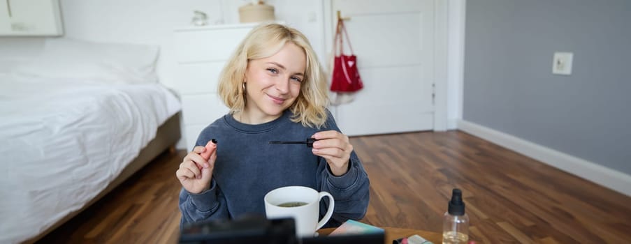 Portrait of cute lifestyle blogger sits in her room in front of video camera, recording for social media account, putting on makeup, reviewing beauty products, holding mascara.