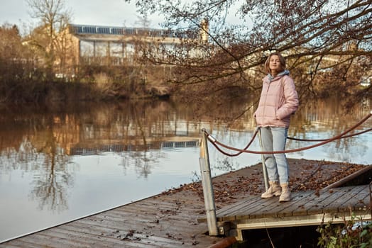 A young woman standing at the shore looking at the river in autumn sunny day. Street view, copy space for text, travel photo. Happy tourist woman on the bank of the river in autumn in warm clothes. Tourists enjoy their vacation, winter season. Romantic look and travel concept. A joyful mood in a Caucasian girl. Winter Wonderland: Enchanting Girl by the Riverside in Autumn.