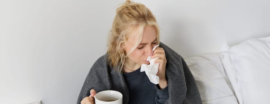 Concept of flu and quarantine. Close up portrait of young woman feeling sick, caught a cold, sneezing and blowing nose in napkin, holding warm tea in hand.