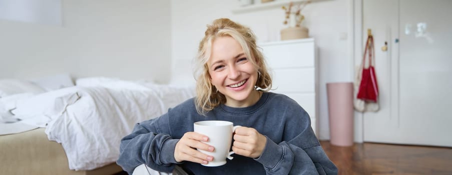 Portrait of young woman sitting on bedroom floor, drinking tea, holding white mug and smiling at camera.