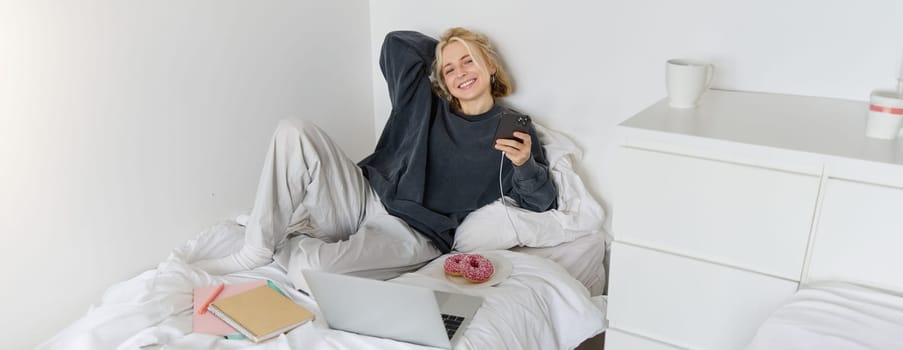 Portrait of young beautiful blond woman, feeling lazy at home, lying in bed with cup of tea, doughnut and laptop, studying remotely or watching movie online.