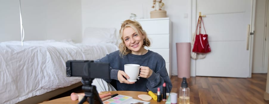 Portrait of young woman, social media influencer, blogger sitting in front of camera with cup of tea and talking, recording video in her bedroom.