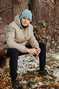 Embrace the tranquility of winter with this captivating image featuring a 17-year-old teen seated on a snowy bench amidst a serene rural park. Sipping tea from a thermos, the young man immerses himself in the enchanting beauty of the snow-covered landscape. This picturesque scene captures the essence of winter relaxation, offering a peaceful moment of solitude and connection with nature during a cozy picnic in the heart of winter.