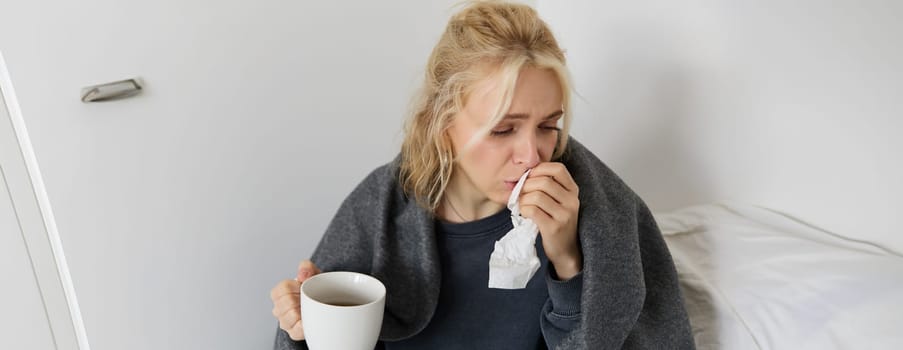 Portrait of woman catching a cold, staying home sick, drinking tea, sneezing in napkin, blowing nose, has covid symptoms.