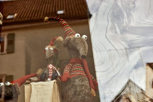 Whimsical New Year's Mouse in Cap and Scarf Stands Behind Store Display. Capture the charm of the holiday season with this delightful image featuring a toy mouse or rat wearing a comical New Year's cap and scarf, playfully standing behind a shop window display. The festive scene adds a touch of whimsy to your creative projects, invoking the spirit of celebration and joy.