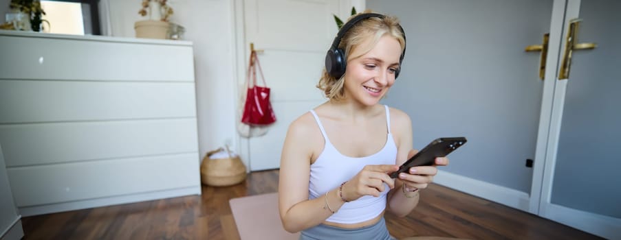 Portrait of young blond woman in headphones, turning on yoga, workout app on smartphone, choosing music on mobile phone application, sitting on rubber mat in room.