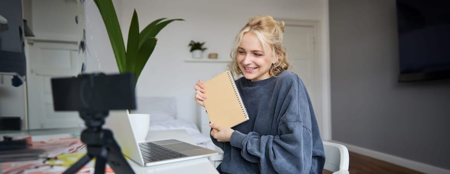 Portrait of cute young woman, teenage girl records video on digital camera, uses laptop to create lifestyle content, shows notebook, reads notes, using computer in her room.